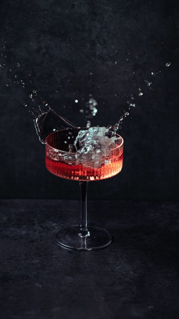 Ein roter Cocktail in einem eleganten Glas, in den gerade Eiswürfel fallen und dabei eine kräftige Spritzbewegung verursachen.